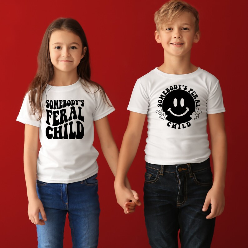 Może przedstawiać: Dwoje dzieci w białych koszulkach z czarnym nadrukiem. Koszulka dziewczynki ma napis "Somebody's Feral Child". Koszulka chłopca ma napis "Somebody's Feral Child" z uśmiechniętą buźką i znakami pokoju. Trzymają się za ręce i stoją przed czerwonym tłem.