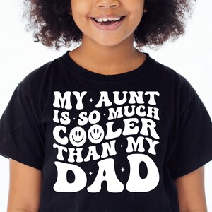 May include: Black t-shirt with the text "MY AUNT IS SO MUCH COOLER THAN MY DAD" in white, retro-style lettering. The word "COOLER" features a smiley face. A fun, casual tee for kids.