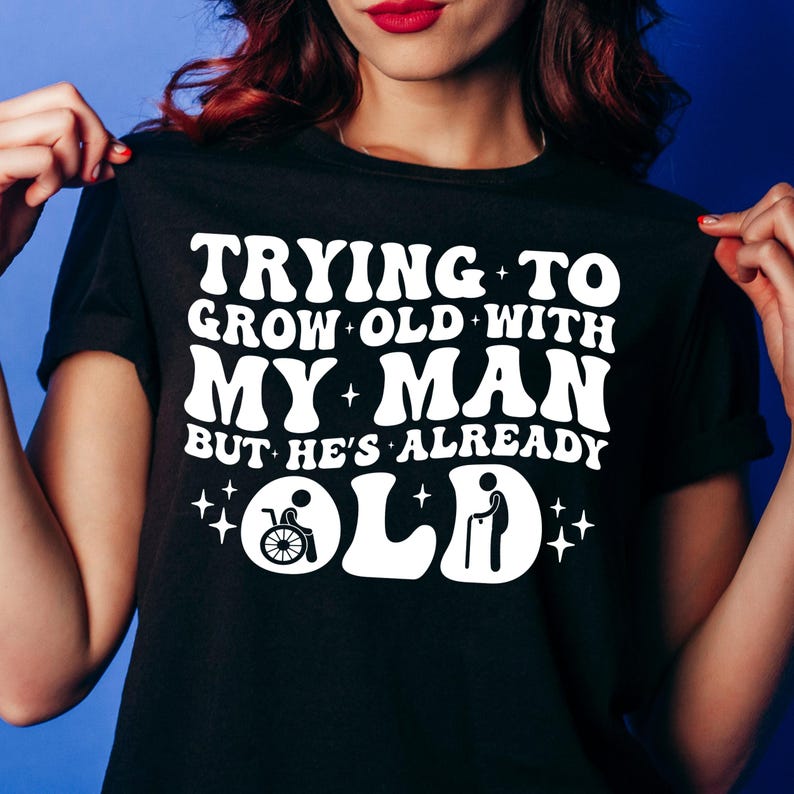 May include: Black t-shirt with white text that reads "Trying to grow old with my man but he's already old". The text is in a retro font style. There are two small white icons below the text, one of a person in a wheelchair and one of a person with a cane.