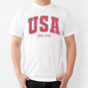 Op de afbeelding: Wit T-shirt met een rode en blauwe vintage-stijl grafische afbeelding met de tekst "USA EST. 1776".