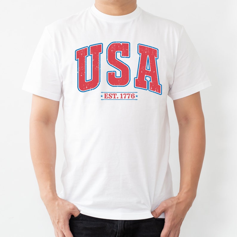Op de afbeelding: Wit T-shirt met een rode en blauwe vintage-stijl grafische afbeelding met de tekst "USA EST. 1776".