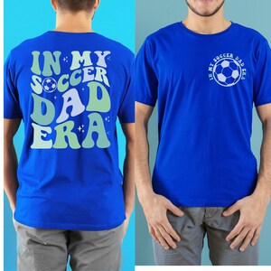 Op de afbeelding: Koningsblauw t-shirt met de tekst "IN MY SOCCER DAD ERA" in een retro lettertype op de rug en een voetbalafbeelding. De voorkant van het shirt heeft de tekst "IN MY SOCCER DAD ERA" rond een voetbalafbeelding.