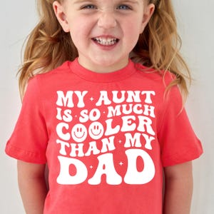 May include: A red t-shirt with the text "MY AUNT IS SO MUCH COOLER THAN MY DAD" in white. The shirt has a crew neck and short sleeves. A young child is wearing the shirt and smiling.