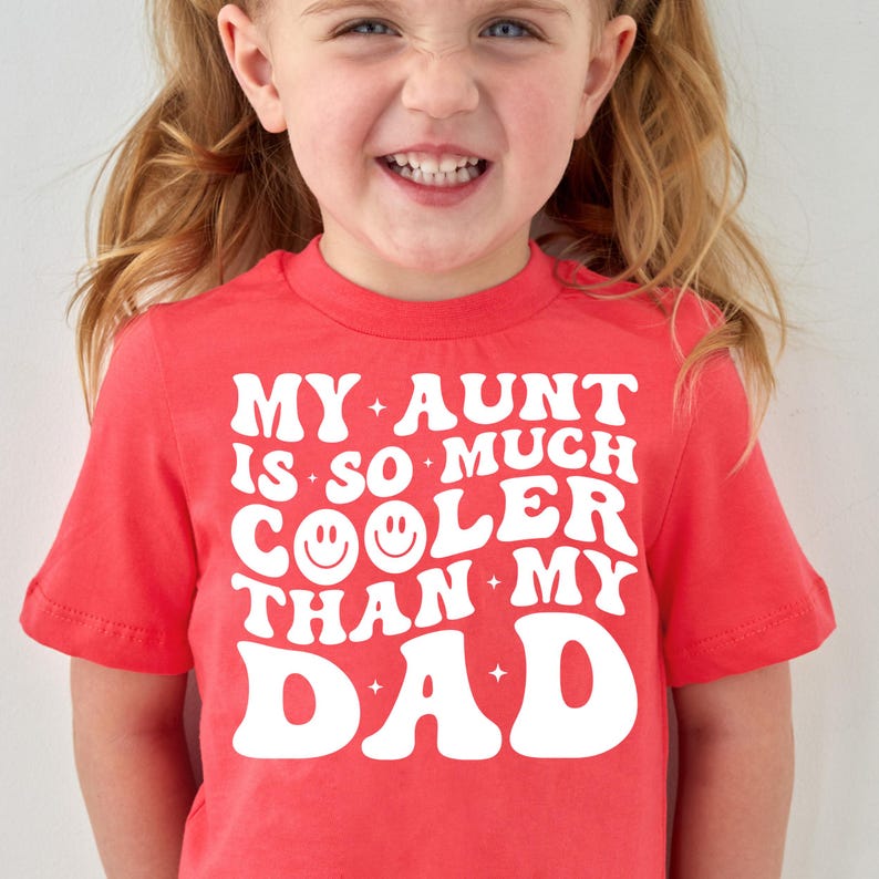 May include: A red t-shirt with the text "MY AUNT IS SO MUCH COOLER THAN MY DAD" in white. The shirt has a crew neck and short sleeves. A young child is wearing the shirt and smiling.