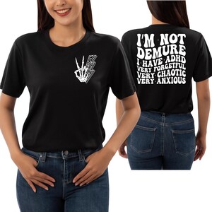 May include: Black t-shirt with a white skeleton hand making a peace sign on the front and the text "I'm not demure I have ADHD very forgetful very chaotic very anxious" on the back.