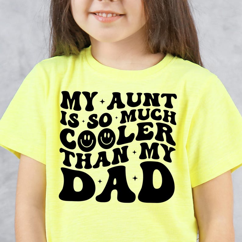 May include: A yellow t-shirt with the text "MY AUNT IS SO MUCH COOLER THAN MY DAD" in black, retro-style lettering. The word "COOLER" features two smiley face emojis. The shirt is worn by a child.