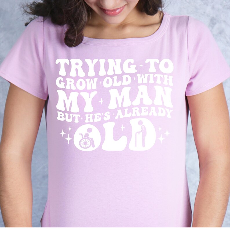 May include: A pink t-shirt with a white graphic that says "Trying to grow old with my man but he's already old". The graphic includes a wheelchair and a cane.