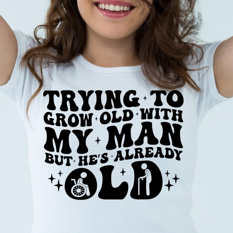 May include: White t-shirt with black text that reads "Trying to grow old with my man but he's already old". The text is in a retro font style. There are two small stick figure icons, one in a wheelchair and one with a cane.