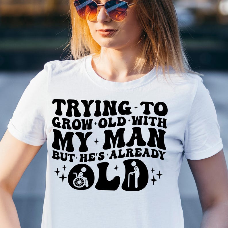 May include: White t-shirt with black text that reads "Trying to grow old with my man but he's already old". The text is in a retro font style. There are two icons below the text, one of a person in a wheelchair and one of a person with a cane.
