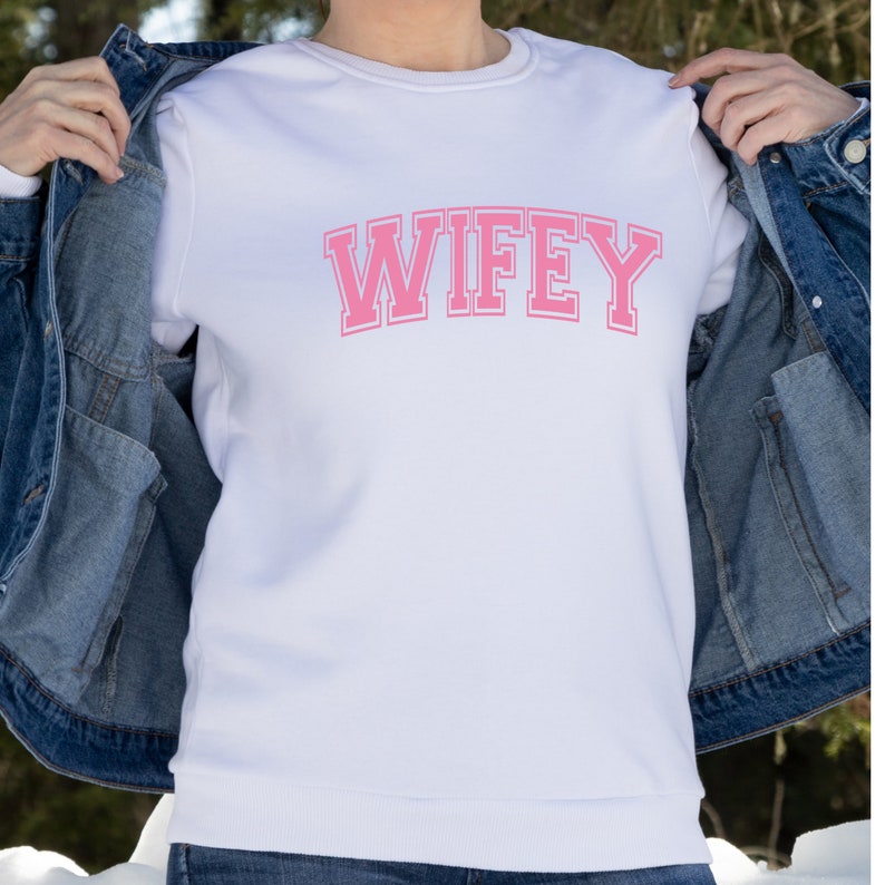 May include: White sweatshirt with the word "WIFEY" in pink block letters across the chest. The sweatshirt has a crew neck and long sleeves. The text is in a collegiate style font.
