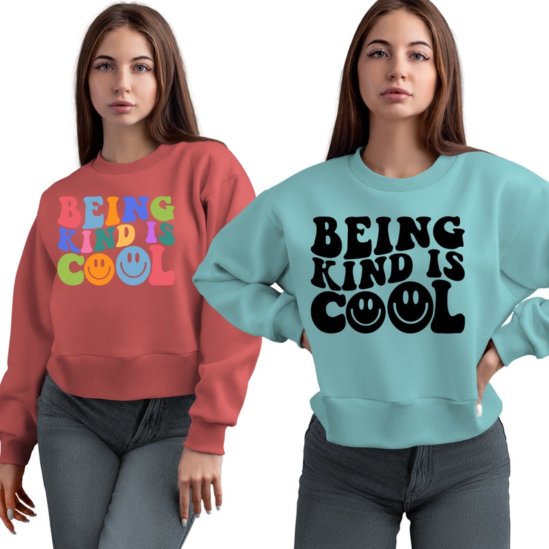 May include: Two women wearing sweatshirts with the text "Being Kind is Cool" in different colors and styles. One sweatshirt is red with colorful letters and smiley faces, the other is blue with black letters and smiley faces.