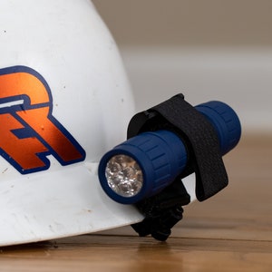 May include: A blue flashlight with a black strap attached to a white hard hat. The flashlight has multiple LED lights and a textured blue body.