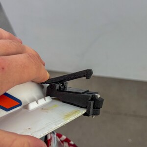 Op de afbeelding: Een zwarte 3D-geprinte klem is bevestigd aan een wit oppervlak met oranje en blauwe graphics. De klem heeft een hendel en wordt door een hand vastgehouden. De achtergrond is een effen wit oppervlak.
