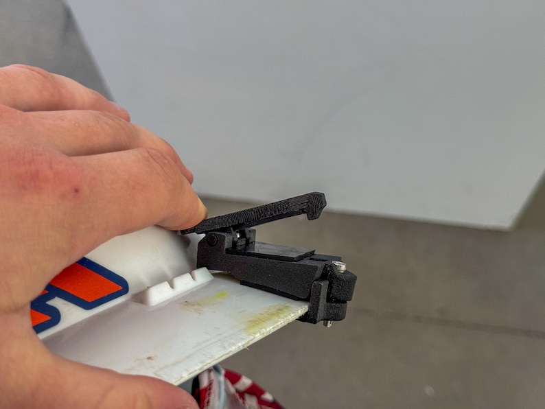 Op de afbeelding: Een zwarte 3D-geprinte klem is bevestigd aan een wit oppervlak met oranje en blauwe graphics. De klem heeft een hendel en wordt door een hand vastgehouden. De achtergrond is een effen wit oppervlak.