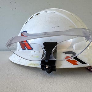 Op de afbeelding: Een witte veiligheidshelm met een heldere veiligheidsbril bevestigd. De helm heeft rode en zwarte afbeeldingen. De bril wordt op zijn plaats gehouden door een zwarte beugel. De helm is waarschijnlijk voor bouw- of industri&euml;le werkzaamheden.