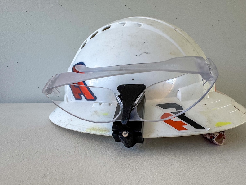 Op de afbeelding: Een witte veiligheidshelm met een heldere veiligheidsbril bevestigd. De helm heeft rode en zwarte afbeeldingen. De bril wordt op zijn plaats gehouden door een zwarte beugel. De helm is waarschijnlijk voor bouw- of industri&euml;le werkzaamheden.