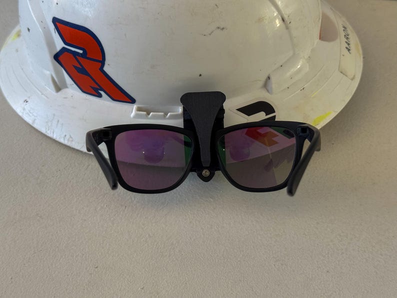 Op de afbeelding: Een witte veiligheidshelm met een zwarte beugel die een zwarte zonnebril met paarse glazen vasthoudt. De helm heeft een rood en blauw logo op de voorkant.