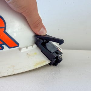 Op de afbeelding: Een zwart clip-on accessoire bevestigd aan een witte veiligheidshelm. De veiligheidshelm heeft een oranje en blauw logo. De clip lijkt van plastic te zijn en is ontworpen om aan de rand van de helm te worden bevestigd.