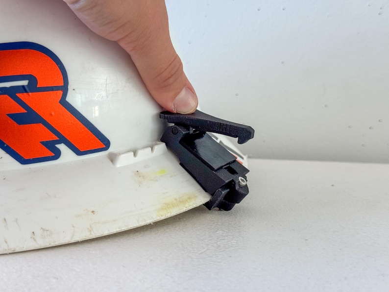 Op de afbeelding: Een zwart clip-on accessoire bevestigd aan een witte veiligheidshelm. De veiligheidshelm heeft een oranje en blauw logo. De clip lijkt van plastic te zijn en is ontworpen om aan de rand van de helm te worden bevestigd.