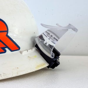 Op de afbeelding: Witte veiligheidshelm met een oranje en blauw logo, voorzien van een transparant plastic vizier. Het vizier is scharnierend voor op- en neerwaartse beweging en biedt oogbescherming. De helm is ontworpen voor bouw of industri&euml;le toepassingen.