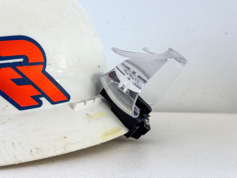 Op de afbeelding: Witte veiligheidshelm met een oranje en blauw logo, voorzien van een transparant plastic vizier. Het vizier is scharnierend voor op- en neerwaartse beweging en biedt oogbescherming. De helm is ontworpen voor bouw of industri&euml;le toepassingen.