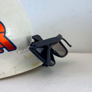 Op de afbeelding: Een witte helm met een oranje en blauw logo, met een zwarte zonnebril aan de zijkant geklikt. De zonnebril heeft een donkere tint en een strak, modern design.