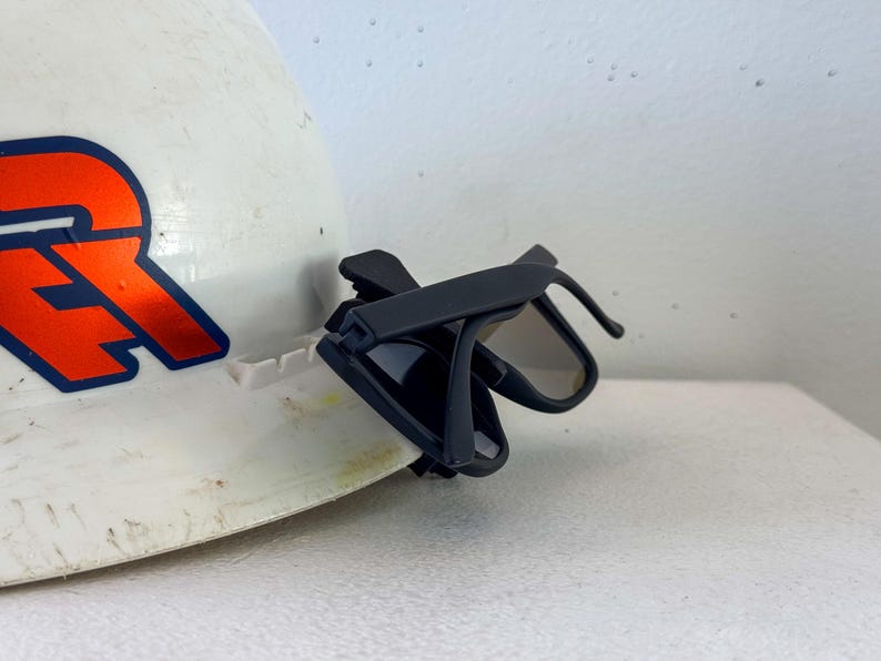 Op de afbeelding: Een witte helm met een oranje en blauw logo, met een zwarte zonnebril aan de zijkant geklikt. De zonnebril heeft een donkere tint en een strak, modern design.