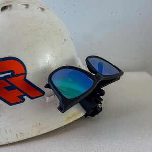 Op de afbeelding: Een witte veiligheidshelm met een rood en blauw logo, gecombineerd met een zwart montuur veiligheidsbril met blauw gespiegelde glazen. De bril is bevestigd aan de helm, wat wijst op een werk- of constructiethema.