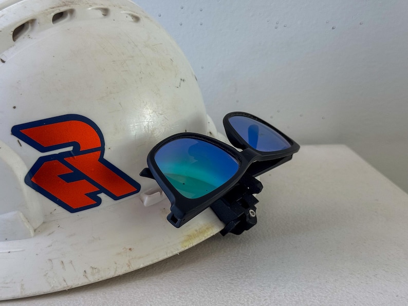 Op de afbeelding: Een witte veiligheidshelm met een rood en blauw logo, gecombineerd met een zwart montuur veiligheidsbril met blauw gespiegelde glazen. De bril is bevestigd aan de helm, wat wijst op een werk- of constructiethema.