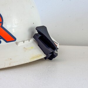 Op de afbeelding: Een witte veiligheidshelm met een oranje en blauw logo. Een zwarte clip is bevestigd aan de rand van de helm. De helm staat op een wit oppervlak.