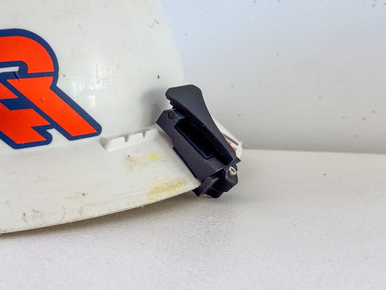 Op de afbeelding: Een witte veiligheidshelm met een oranje en blauw logo. Een zwarte clip is bevestigd aan de rand van de helm. De helm staat op een wit oppervlak.
