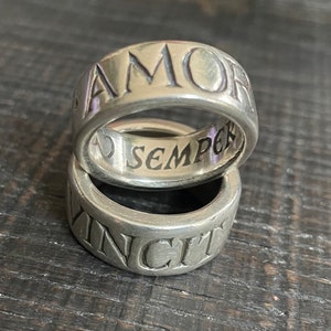May include: Two silver rings stacked on top of each other. The top ring has the word "AMOR" engraved on the outside and the word "SEMPER" engraved on the inside. The bottom ring has the word "VINCIT" engraved on the outside.