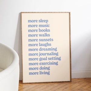 May include: A framed print with a beige background and blue text that reads "more sleep, more music, more books, more walks, more sunsets, more laughs, more dreaming, more journaling, more goal setting, more exercising, more doing, more living."