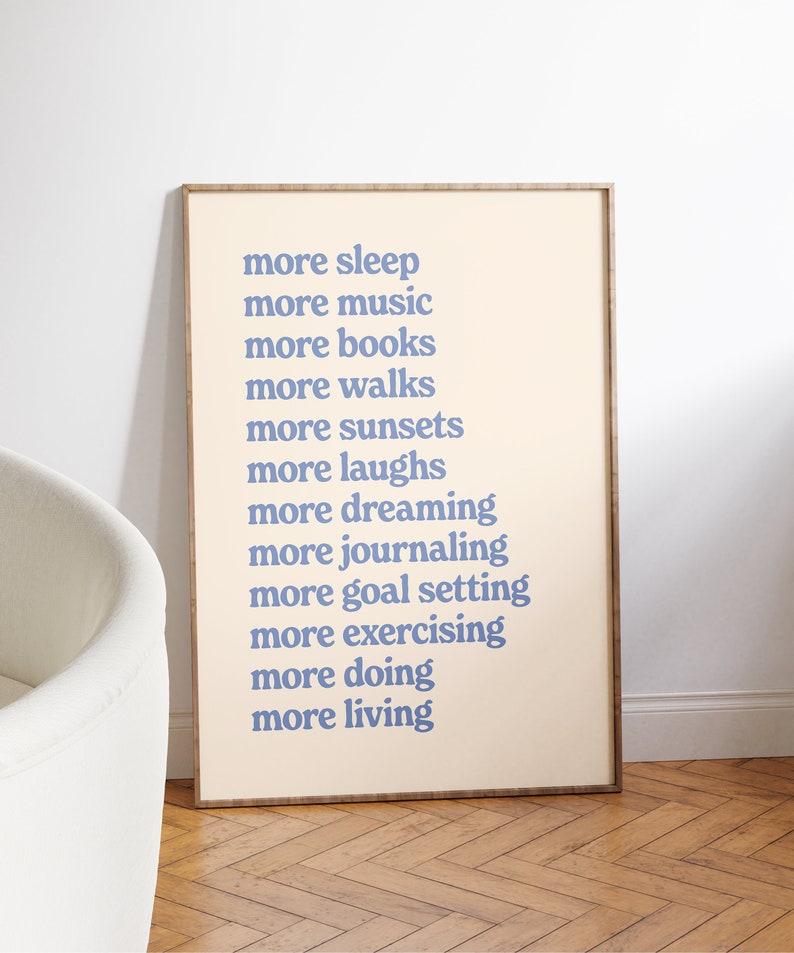 May include: A framed print with a beige background and blue text that reads "more sleep, more music, more books, more walks, more sunsets, more laughs, more dreaming, more journaling, more goal setting, more exercising, more doing, more living."