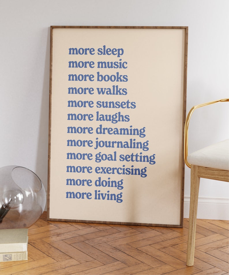 May include: A beige framed print with blue text that reads "more sleep, more music, more books, more walks, more sunsets, more laughs, more dreaming, more journaling, more goal setting, more exercising, more doing, more living."