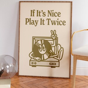 May include: A framed print with a beige background and gold text that reads "If It's Nice Play It Twice". A cartoon illustration of a record player with a record that has a face and is giving the peace sign is in the center of the print.