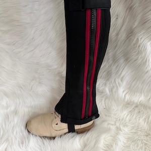 May include: Black leg warmers with a red stripe and a zipper detail. The leg warmers are worn over a pair of beige shoes. The leg warmers have a buckle closure at the top and bottom.