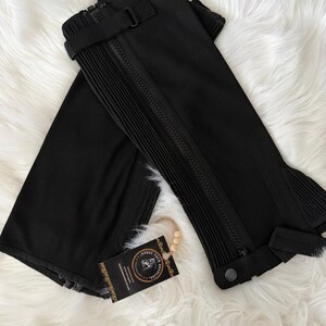 May include: Black riding half chaps with zippers, buckles, and pleated detailing. The chaps are made of a durable material and are designed to protect the rider's legs. A tag with the text "Horse Tack Supplies" is attached.