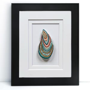 May include: A framed artwork featuring a colorful, embroidered shell design. The shell is in shades of teal, orange, and gold, set against a white mat and framed in a black rectangular frame. The artwork is a decorative piece.