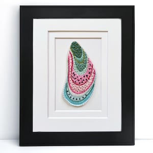 May include: Framed artwork featuring a colorful, embroidered oyster shell. The shell is primarily pink, green, and blue, with intricate stitching and a textured appearance. The artwork is set against a white background within a black frame.