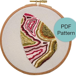 May include: An embroidery hoop with a colorful, abstract design resembling a geode. The design features shades of red, pink, green, and brown, stitched onto a neutral fabric. A teal circle with the text "PDF Pattern" is on the right.