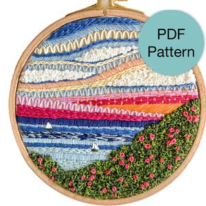 May include: A circular wooden embroidery hoop containing a landscape needlework design featuring a blue sky, pink and orange hills, a blue sea with two small sailboats, and a foreground of green bushes with pink flowers. A teal circle says PDF Pattern.