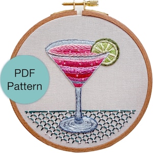 May include: Embroidered cocktail design in a wooden hoop. The drink is pink with a lime garnish. The text "PDF Pattern" is on a light blue circle. The background is a light beige fabric.