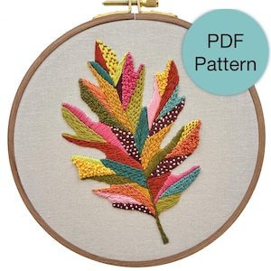 May include: A colorful embroidery pattern of a stylized leaf in a wooden embroidery hoop. The pattern is made up of various shades of green, yellow, orange, pink, and red. The text "PDF Pattern" is visible in a light blue circle.