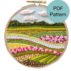 May include: Embroidered hoop art depicting a landscape scene with a blue sky, white clouds, green hills, yellow fields, and pink flowers. The hoop is made of wood and has a natural finish. PDF Pattern.