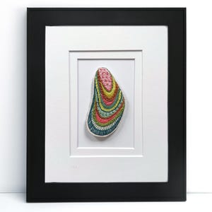 May include: A framed artwork featuring a colorful, abstract design resembling a shell or seed. The artwork is primarily composed of pink, yellow, green, and blue hues, with intricate patterns. The frame is black, and the artwork is matted in white.