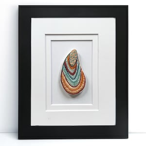 May include: A framed artwork featuring a colorful, abstract shell design. The shell is composed of layered, textured materials in shades of orange, teal, and gold. The artwork is set within a white mat and a black frame.