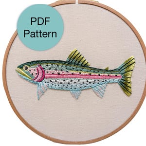 May include: An embroidered rainbow trout design in a wooden hoop. The fish is in shades of green, blue, pink, and yellow. The text "PDF Pattern" is in a light blue circle in the upper left corner. The background is a light beige fabric.