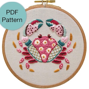Peut inclure: Broderie d'un crabe dans un cercle en bois. Le crabe est brodé avec des fils roses, turquoise et jaunes. Le texte "PDF Pattern" est dans un cercle turquoise en haut à gauche. La broderie est sur un tissu beige clair.