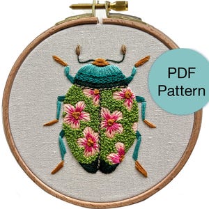 Puede incluir: Diseño de insecto bordado en un aro de madera. El insecto presenta una cabeza y patas turquesas, un cuerpo verde con acentos de flores rosas y antenas marrones. El texto "PDF Pattern" está en un círculo turquesa.
